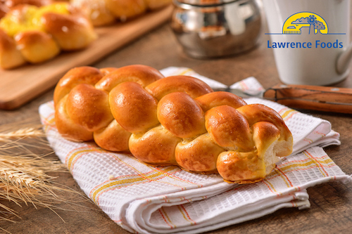 Golden-brown braided brioche loaf on a rustic breakfast table with wheat accents.
